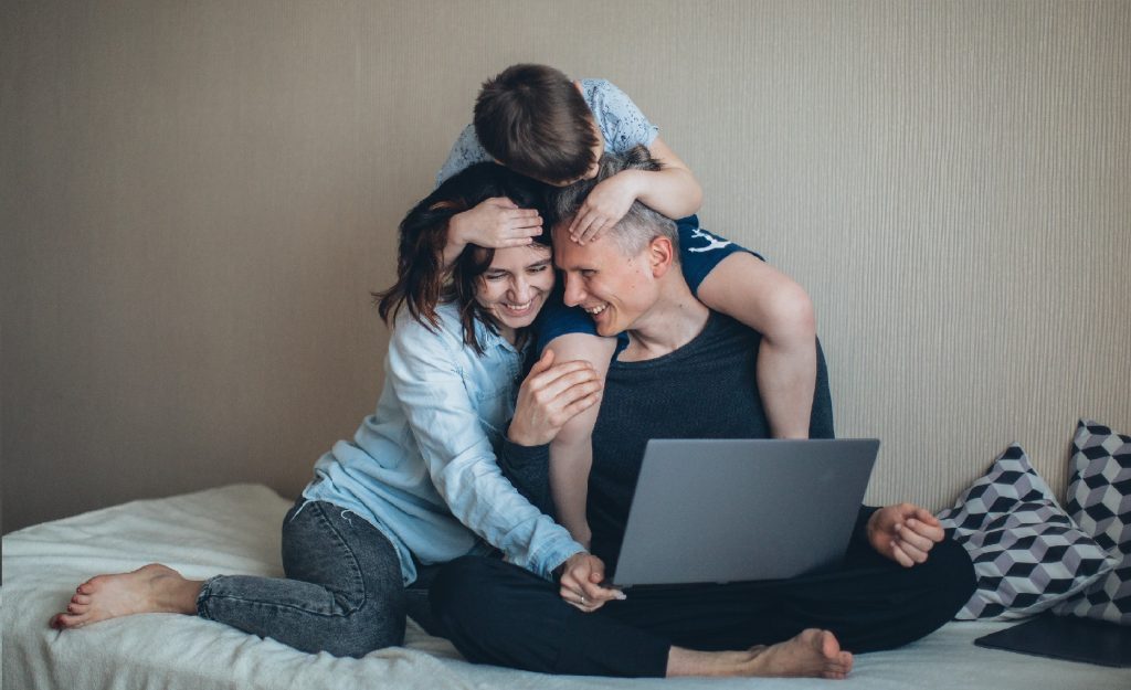 Parents sitting on a bed and looking at a laptop while their son is hugging them.